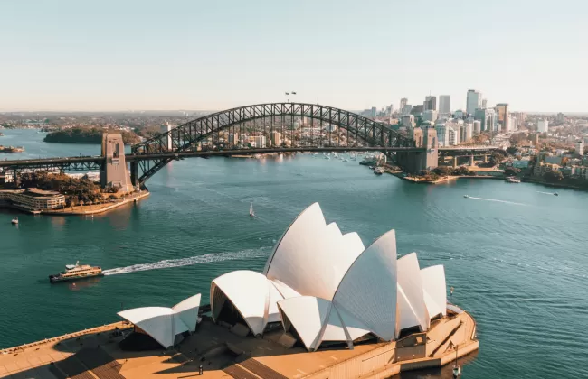 Aerial view of Sydney Harbour with the Sydney Opera House and Harbour Bridge under a clear sky.