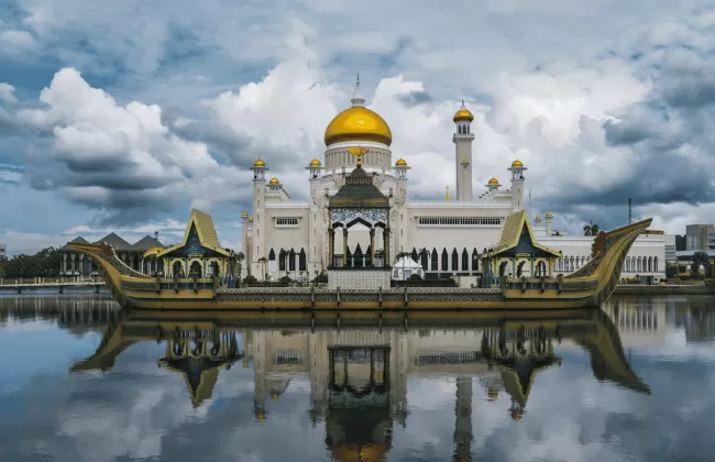 Grand mosque with golden dome and white minarets reflected in calm water under a dramatic cloudy sky in Brunei.