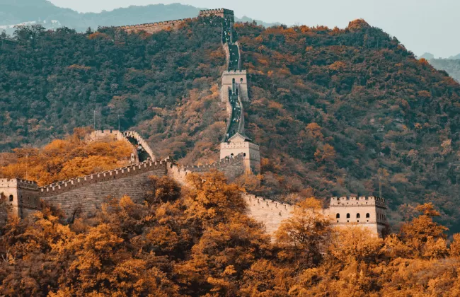 Section of the Great Wall of China winding through forested mountains with autumn-colored trees.