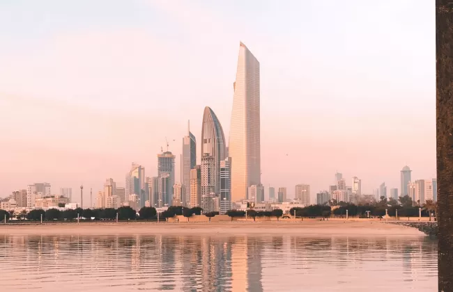 A modern skyline of Kuwait City with reflective buildings and a pink sky at sunset.