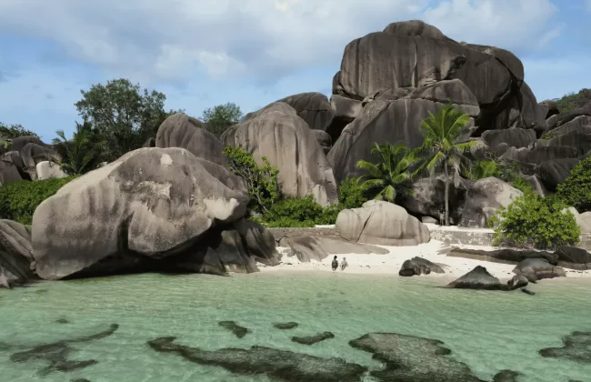 Granite boulders and palm trees by turquoise waters and sandy beach.