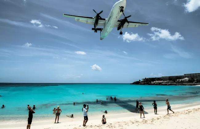 A plane flying low over a turquoise beach with people below.