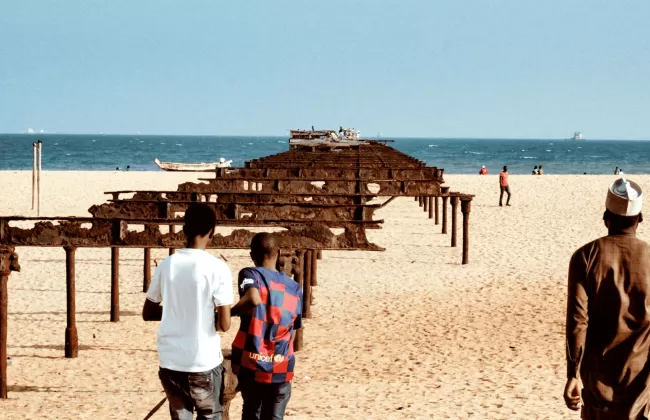 People walking along a sandy beach with rusted pier structures.