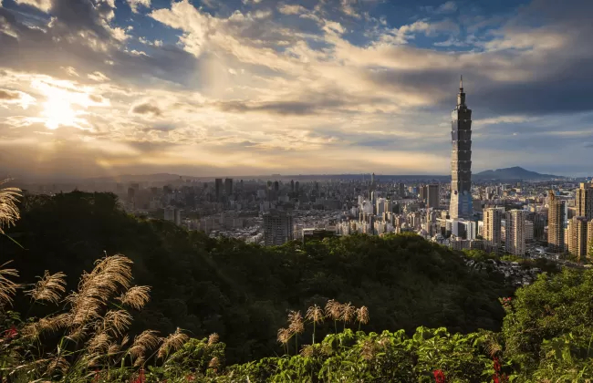 City skyline with Taipei 101 skyscraper framed by grassy hills at sunset.