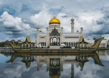 Grand mosque with golden dome and white minarets reflected in calm water under a dramatic cloudy sky in Brunei.