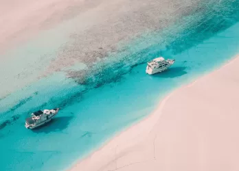 Aerial view of two boats floating over vibrant turquoise water and pink sand in the Bahamas.