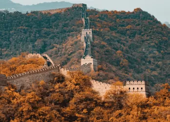Section of the Great Wall of China winding through forested mountains with autumn-colored trees.