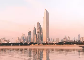 A modern skyline of Kuwait City with reflective buildings and a pink sky at sunset.