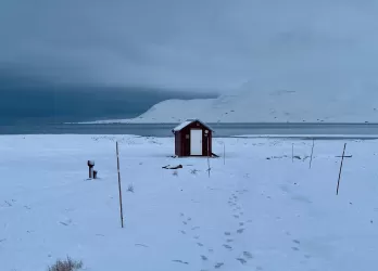 Snow-covered landscape with a small red hut and mountains in the distance.