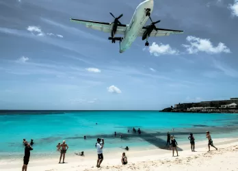 A plane flying low over a turquoise beach with people below.