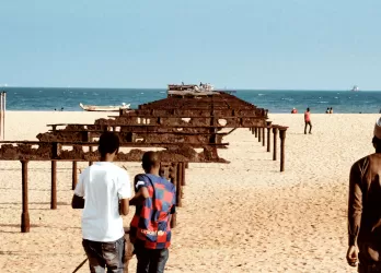 People walking along a sandy beach with rusted pier structures.