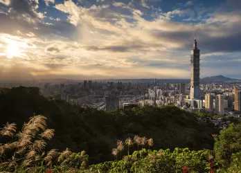 City skyline with Taipei 101 skyscraper framed by grassy hills at sunset.